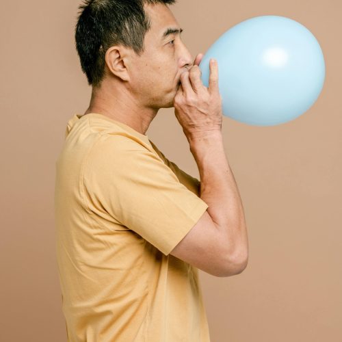 Side view of man in casual t-shirt blowing a blue balloon with a beige background.